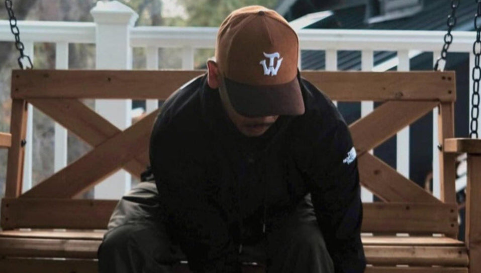 Model wearing a brown structure baseball snapback hat at a cabin in the mountains, sitting on a swinging wooden chair on the porch.  Model is looking down and motioning as if he is tying his shoes